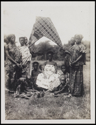 Female, possibly chief's wife, seated under a pair of fans, surrounded by 10 women and children all wearing body cloths or skirts and blouses