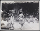 Back view of priest before a group seated under a thatched shelter with a man carrying a bowl and another a stool on left foreground