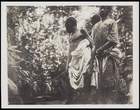 3 males standing in the forest before seated women and children