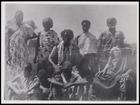5 males seated in the foreground and 4 standing in the background, all wearing body cloths