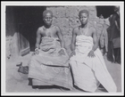 2 females in decorative body cloths, bracelets and 1 with a necklace, seated in front of a mud house