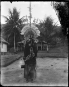 back view of man holding a drum, wearing a large elaborate feather headdress (oa oa ?) standing on dancing ground ?