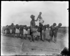 young child, being supported by a man wearing a striped cloth skirt, walking along the backs of a line of other children standing on a beach, part of a game called cuscus