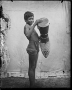 Boy holding large elaborately carved drum, standing in front of white cloth backdrop