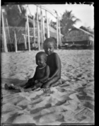 Two small children sitting in the sand in front of houses