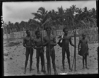 four young men, holding bows and arrows, one carrying a large string bag on a wooden pole, and a young boy carrying a large string bag, all standing on a beach with piles of cut timber (?) in background