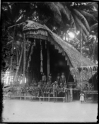large elaborately decorated man's house with group of men and children sitting on front platform, man with cloth over his shoulder appears in Dauncey's Papuan Pictures as a widower
