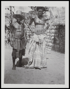 two men, Kabay and Shamba (on left ?), one wearing a European hat ?, standing outside a hut