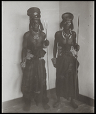 two men, one with paint around his eyes, wearing headdresses made of circular metal plate, holding spears (one with two points) standing in corner of European style room