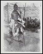 bambi of Mushenge holding wooden staff, seated crowd in the background (and palace walls ?)