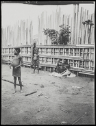 child standing in front of wooden stockade and man sitting by stockade with lengths of raffia ? used for weaving ?