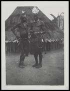 two men wearing headresses standing outside conical thatched hut, man on the left is a 