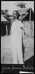 woman wearing cloth dress, with her arms behind her head, standing outside a hut