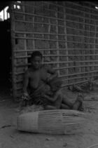 A woman and child sitting outside a wooden building, weaving a type of basket.
