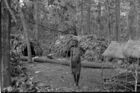 A woman stands facing the camera in the middle of a settlement in a clearing in the forest.