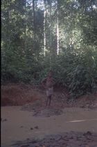 A youth wearing shorts standing on a raised mud bank at the edge of a muddy river or water hole in the forest.