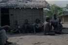 Seven men and youths sat on the ground outside a building made of wooden poles and mud with a thatched roof.