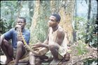 Two men, Adamo and Mamadou, sat on the ground in front of the base of a thick tree trunk.